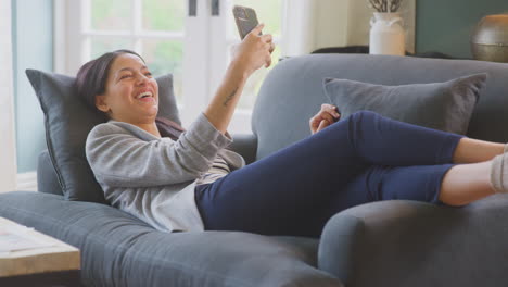 mujer trabajando desde casa tomando un descanso acostada en el sofá transmitiendo una película al teléfono móvil