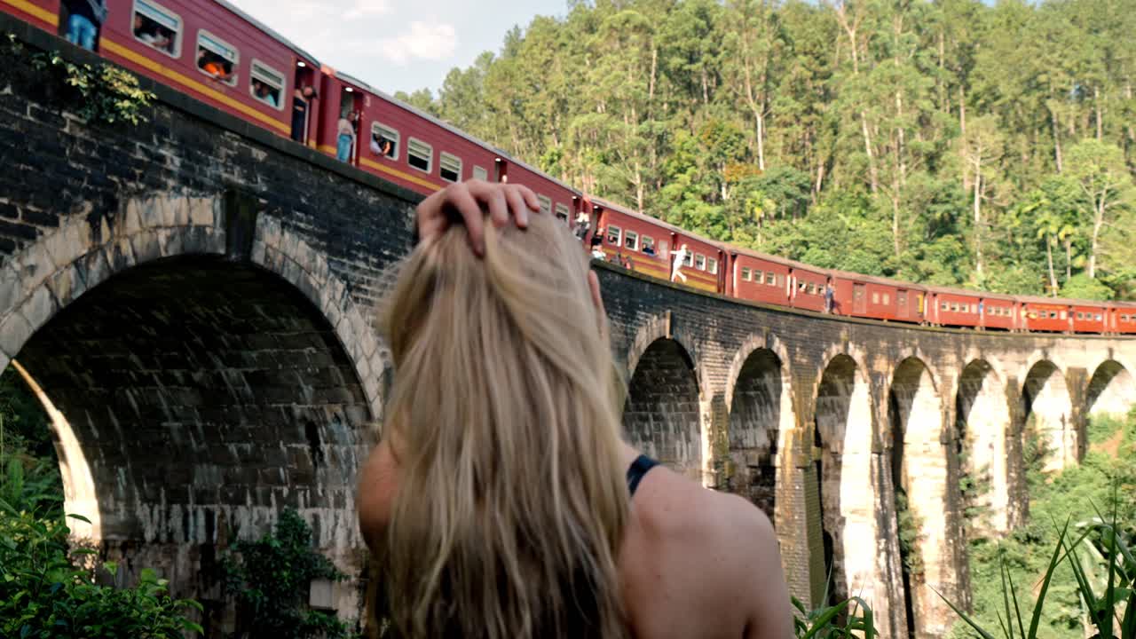 A young woman stands at a viewpoint near the Nine Arch Bridge in Ella, Sri Lanka, gazing at a colorful train as it moves across the historic stone arches.