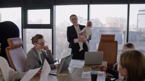 Confident-brunette-businesswoman-with-glasses-in-a-business-suit-holds-a-small-infant-child-in-her-arms-and-communicates-with-her-colleagues-about-an-idea-and-plan-for-work.-During-a-meeting-at-the-table-in-the-office