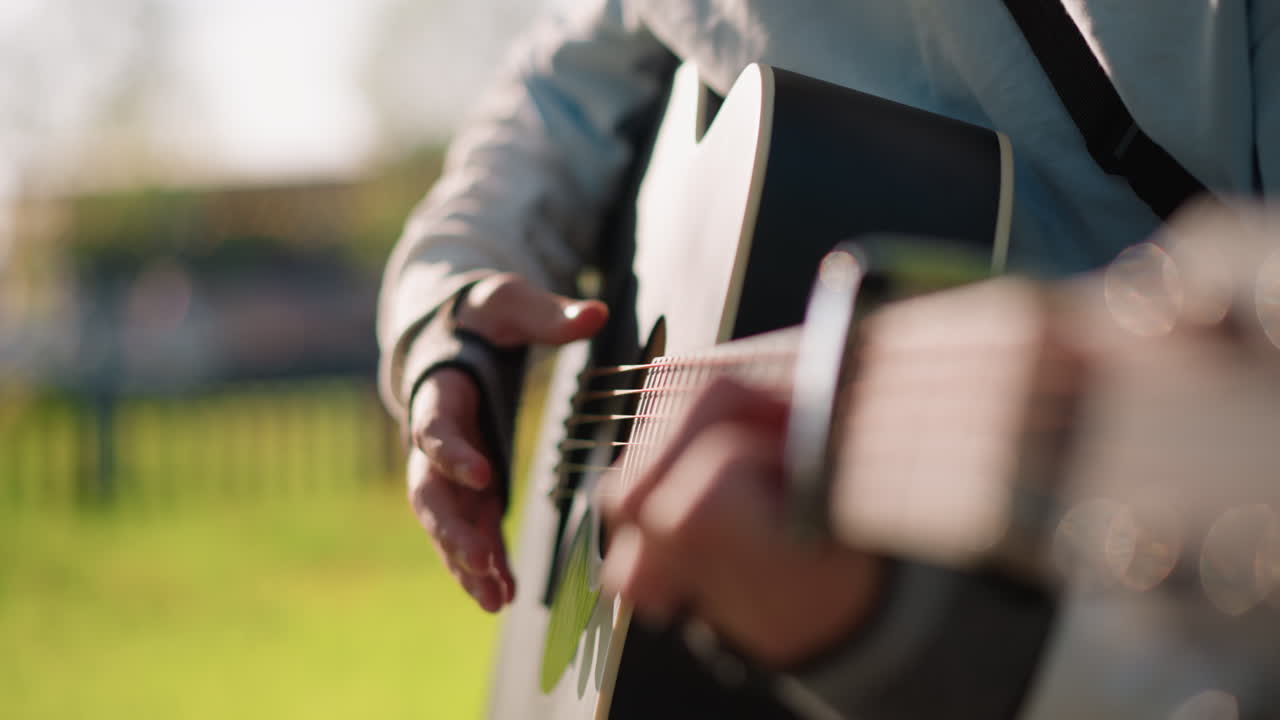 Closeup hands strumming acoustic guitar, outdoor park session with warm sunlight and creamy bokeh, capo clamped near nut, ring on finger, casual jacket sleeve, intimate fingerstyle playing, shallow