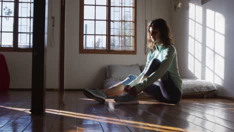 happy mixed race woman preparing for workout, putting on shoes sitting on floor in sunny bedroom