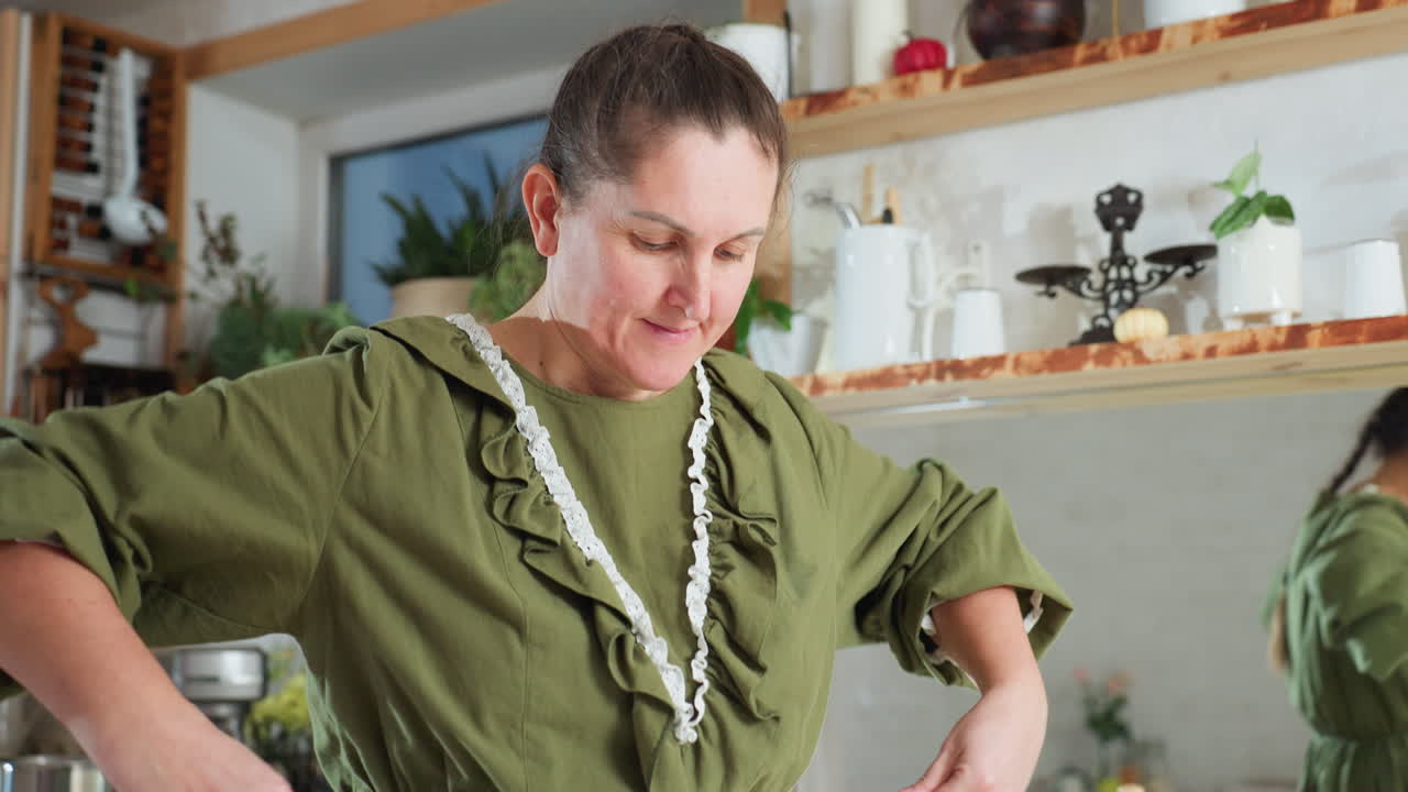 White woman in green dress picks up white apron and begins to tie it around waist in cozy, plant-filled kitchen, captured in warm daylight with mirror reflection