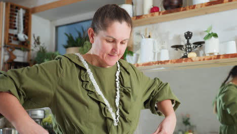 white woman in green dress picks up white apron and begins to tie it around waist in cozy, plant-filled kitchen, captured in warm daylight with mirror reflection