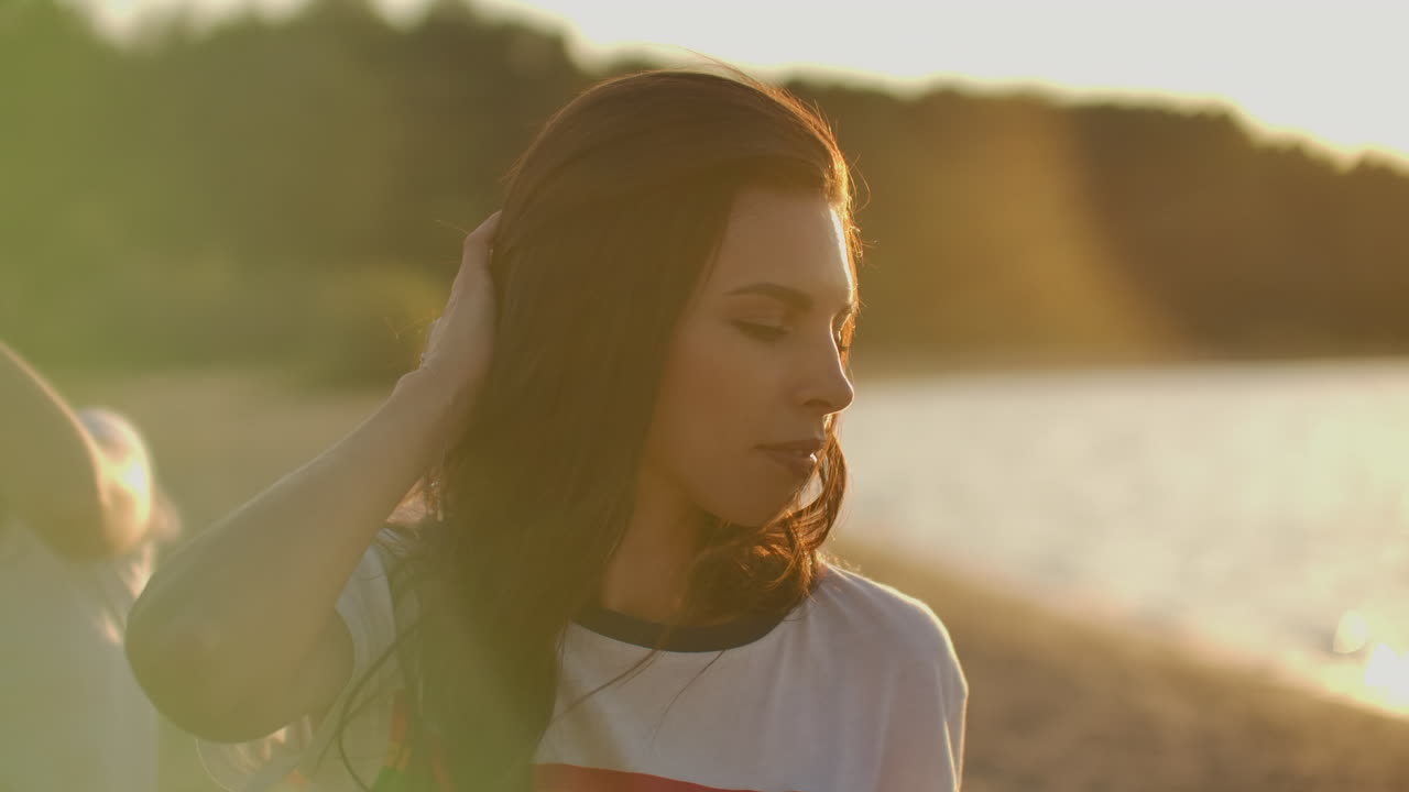 una chica libre con el cabello largo y oscuro en una camiseta blanca corta está bailando en la fiesta de la playa con sus amigos. ella sonríe y toca su cabello y disfruta del tiempo de la fiesta al aire libre.