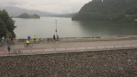 aerial view, orbit style, of a group of cyclists standing over the dam at colibita lake while one cyclist peddles along, during tura cu copaci race