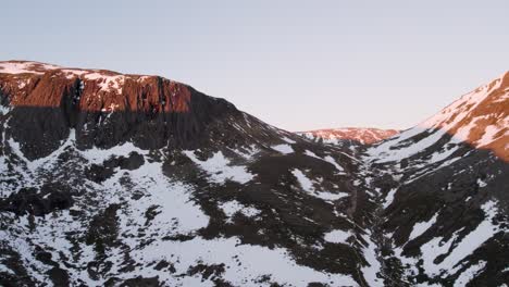 Imágenes-Aéreas-De-Drones-Panorámicos-Para-Revelar-Montañas-Y-Acantilados-Escarpados-Y-Dramáticos-En-Un-Paisaje-Invernal-Con-Nieve,-Rocas-Y-Rocas-Al-Amanecer-En-Una-Parte-Remota-Del-Parque-Nacional-De-Cairngorms,-Escocia