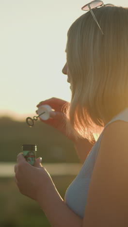 mujer con una camiseta sin mangas azul con gafas de sol en la cabeza soplando burbujas durante la hora dorada, la luz del sol iluminando su cabello, con un fondo natural borroso