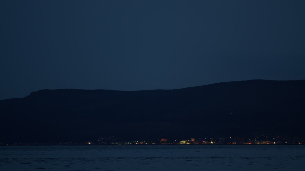 바가의 표면을 가로질러 물의 파동이 울리고 산 아래의 집에 불빛이 켜져 있습니다.