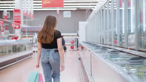 young lady walking through well-lit mall holding shopping bags, looking around while sleek cabinet reflections and hanging signpost are visible, someone ahead of her in the distance