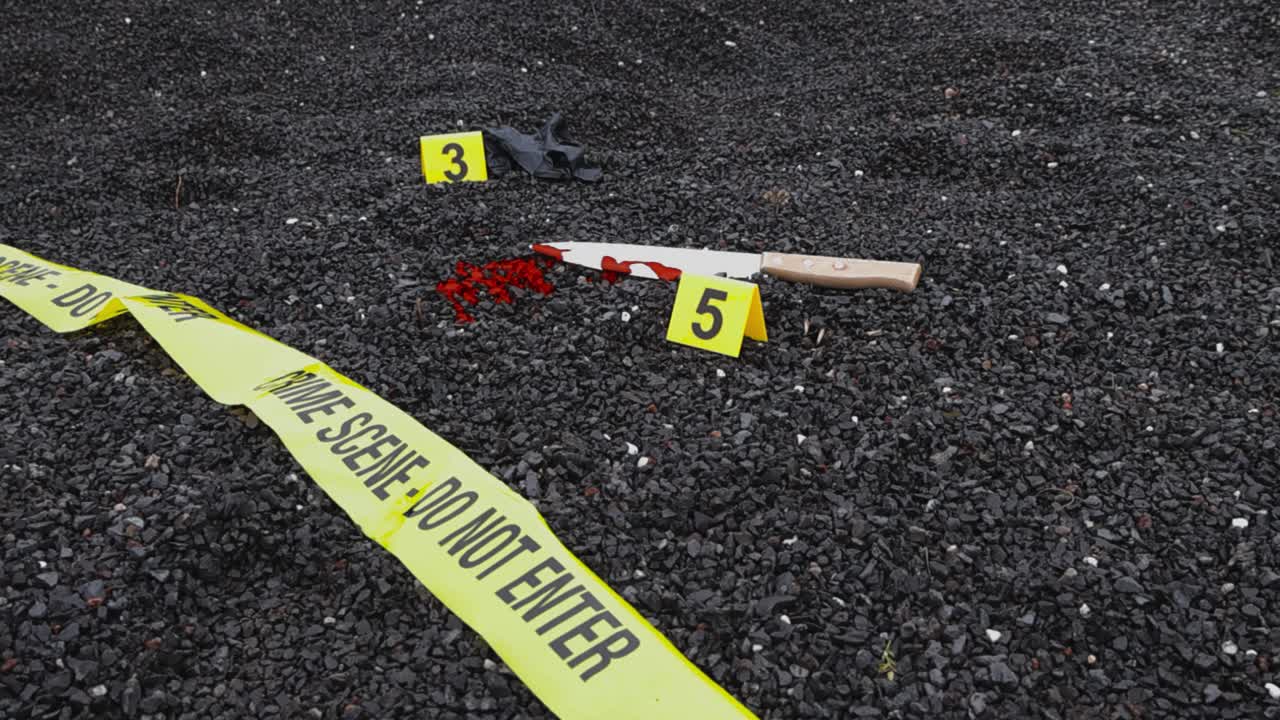 Low angle ascending view reveals forensic evidence markers placed next to discarded rubber glove, blood stained knife and spatter on dark gravel ground. Fallen crime scene tape lies across the terrain