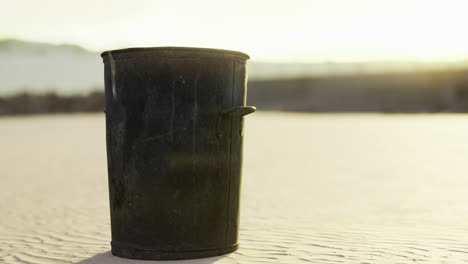 black trash can standing alone on a sandy beach at sunset