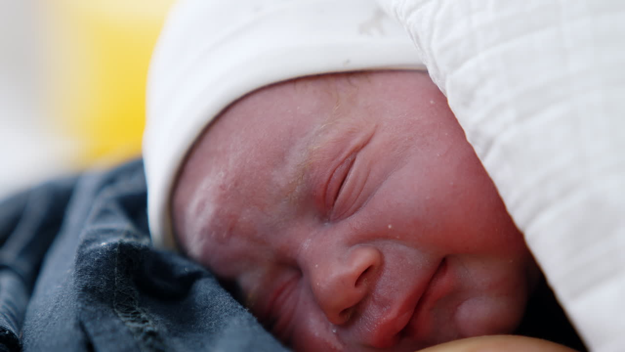 Face of a newborn right after birth. Tiny baby in white cap wants to cry and blinking eyes. Close up.