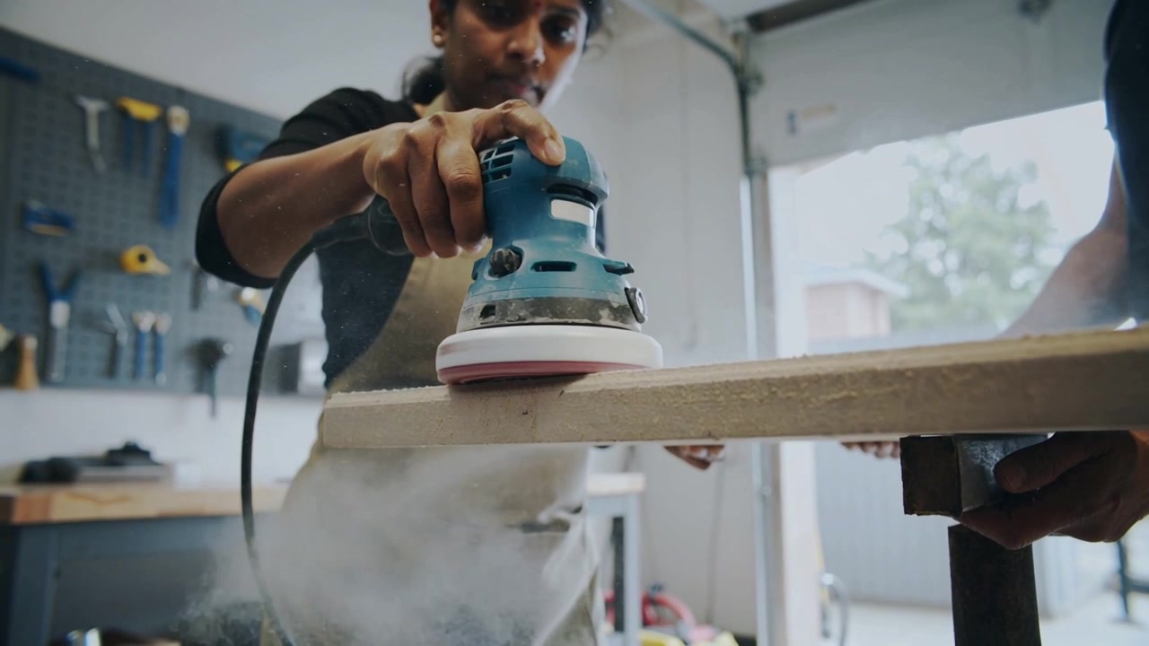 Placing sander, worker in tan apron using blue sander on shop bench smoothing board, helper holding