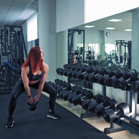 sportswoman in black top and leggings swinging hard kettlebell in fitness club. female athlete doing workout and looking at the mirror near rows of dumbbells.