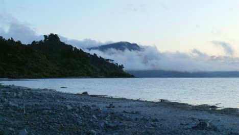 A-breathtaking-long-shot-captures-the-enchanting-scene-of-billowing-clouds-gracefully-enveloping-a-mountain-on-the-shores-of-a-serene-lake,-painting-a-tranquil-and-majestic-natural-panorama