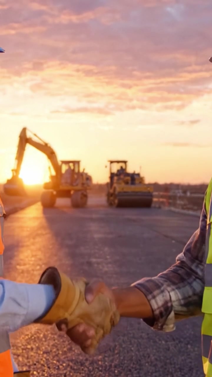Construction Workers Collaborating at Sunset: Two Professionals Discussing Project Progress Amid Heavy Machinery on an Expansive Paved Road