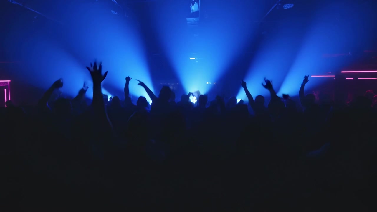 Folla densa che reagisce all'illuminazione, alzando le mani sul palco di un club con faretti blu e LED magenta.