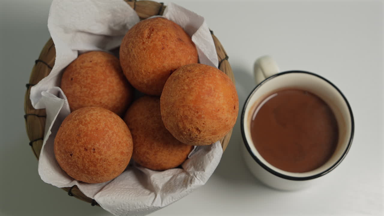 Savor a close-up view of golden, crispy buñuelos in a woven basket beside a steaming mug of hot chocolate. Bright, inviting lighting enhances the warm and cozy mood