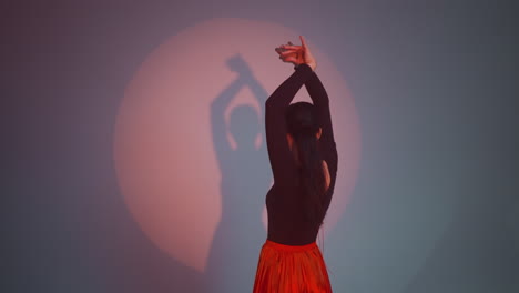 a lady in a black and red gown stands facing the wall, moving her body and hands slowly in a dancing manner against a grey background