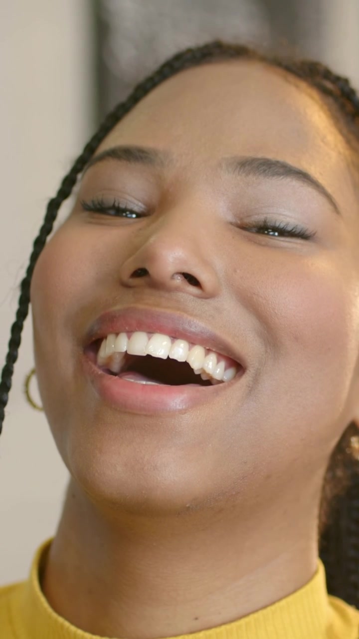Vídeo vertical: Mulher sorrindo no estúdio para a câmera, caindo na risada e inclinando a cabeça, de blusa amarela.