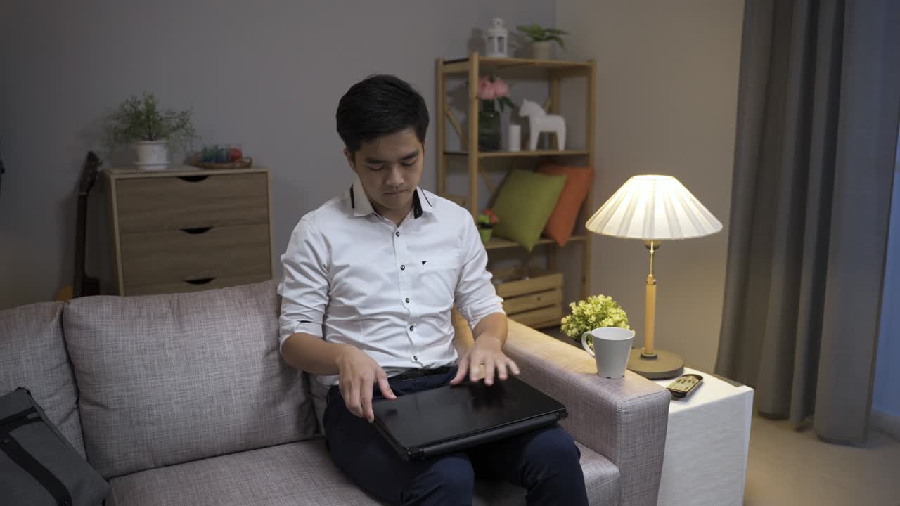 커피를 들고 거실에 들어오는 아시아인 사업가는 소파에 앉아서 노트북을 열고 저녁에 집에서 오버타임을 시작합니다.