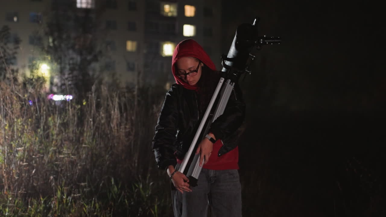 amateur astronomer adjusts telescope at night in dimly lit urban field wearing red hoodie, tripodmounted refractor, apartment windows glowing in background, quiet atmosphere of curiosity