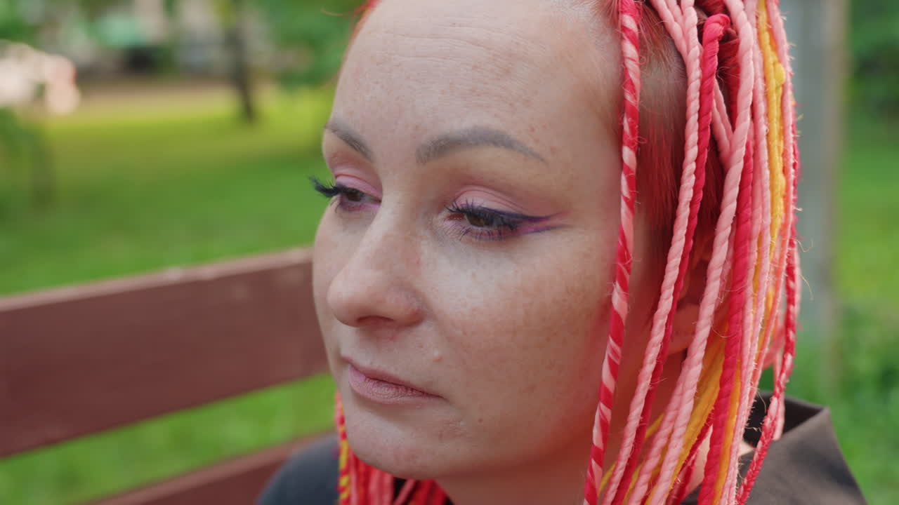 생각에 잠긴 채 공원에 있는 다채로운 머리 색깔의 여성, 활기찬 머리카락을 가진 슬픈 여성이 야외에서 안경을 고쳐 쓰고, 염색한 머리카락을 가진 감성적인 여성이 공원에서 안경을 벗으며 잠시 멈춰 서 있어요.