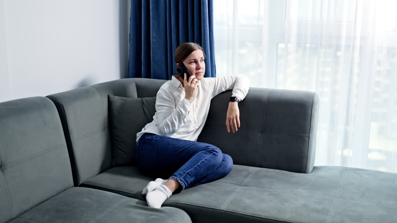 Bella donna in jeans e camicia seduta sul divano a casa che parla al telefono. Giovane donna che lavora online al telefono stando a casa. Lavoro da remoto online.