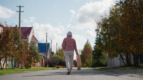 back view of woman in pink jacket and gray sweatpants walks away with paper bag swaying down residential street, surrounded by vibrant autumn foliage and quaint houses under bright skies