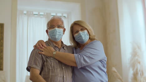 pareja de ancianos con máscara médica de pie en casa y abrazándose