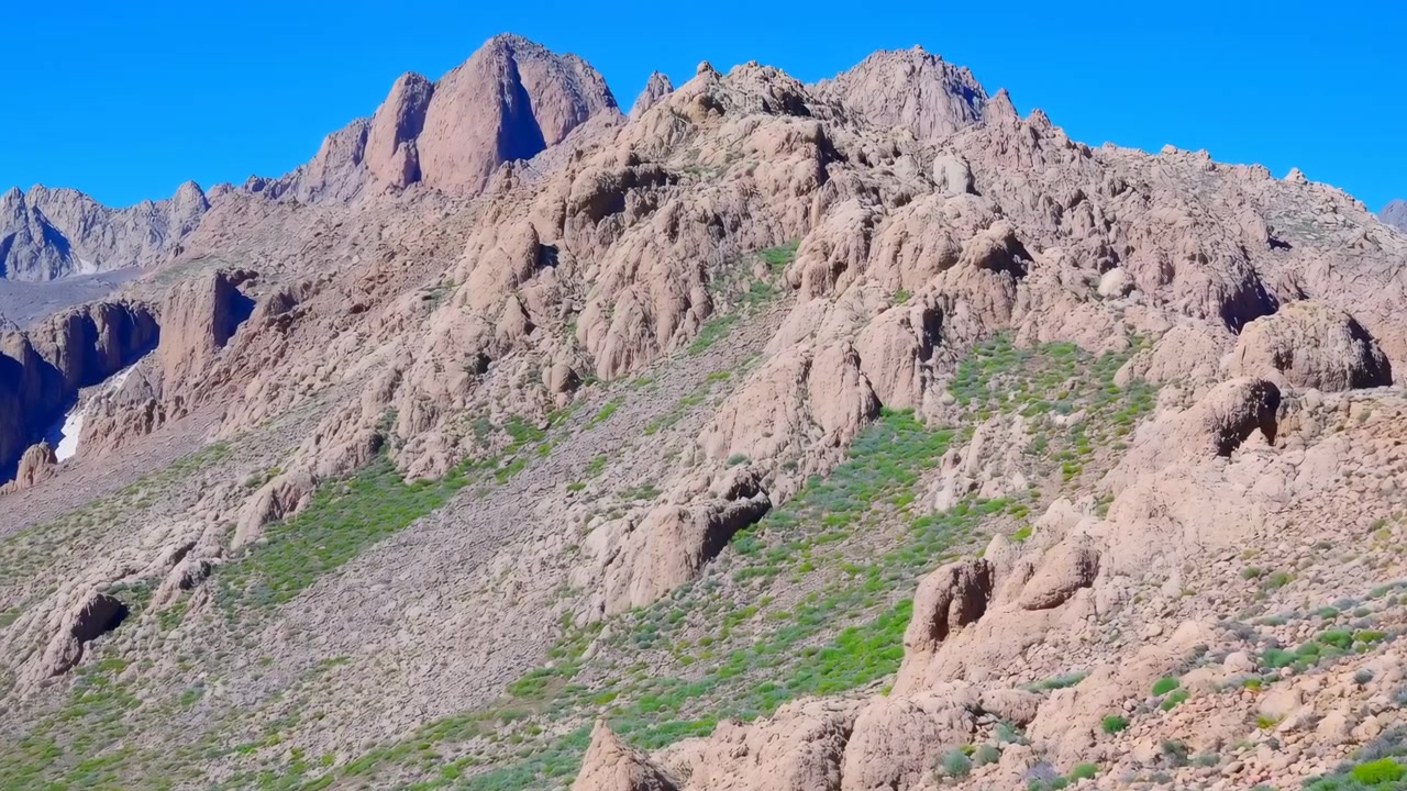 Ampio paesaggio naturale che mostra un ripido pendio roccioso con scogliere frastagliate color beige, arbusti verdi sparsi e formazioni rocciose illuminate dal sole sotto un cielo limpido e luminoso