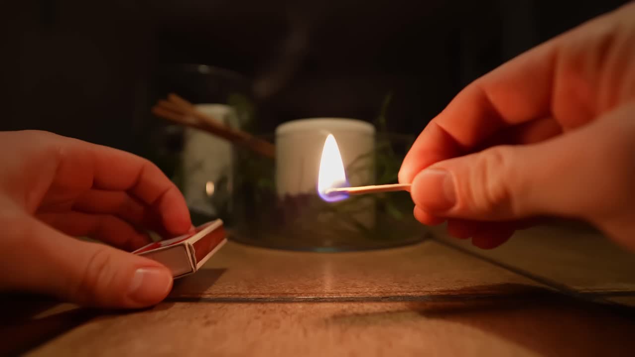 Tangan menyalakan korek api dari kotak korek untuk menyalakan lilin, close-up pada nyala api hangat dan suasana rumah yang nyaman. Sempurna untuk musim dingin, Natal, Tahun Baru, ritual tenang, dan b-roll gaya hidup