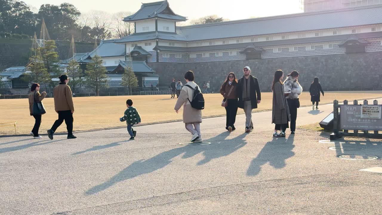 夕暮れ時、舗装された道に長い影が伸びる金沢城の敷地を散策する訪問者たち。
