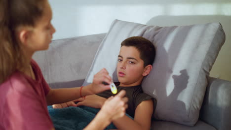 rear view girl with ponytail and maroon shirt gently removes thermometer from mouth of sick boy lying on couch under blanket showing care concern and responsibility in quiet softly lit indoor home