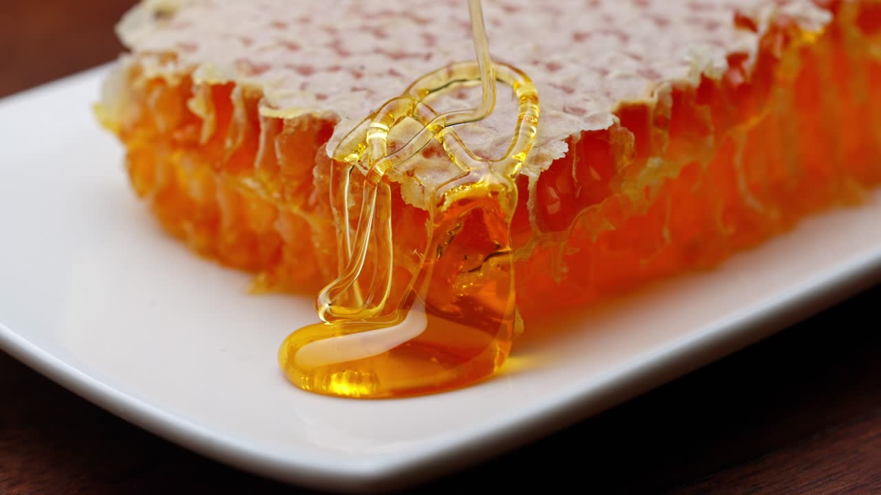 Gocce di miele dorato e denso che colano lentamente su celle di favo fresche in un macro primo piano alimentare pulito, pioggia naturale fresca