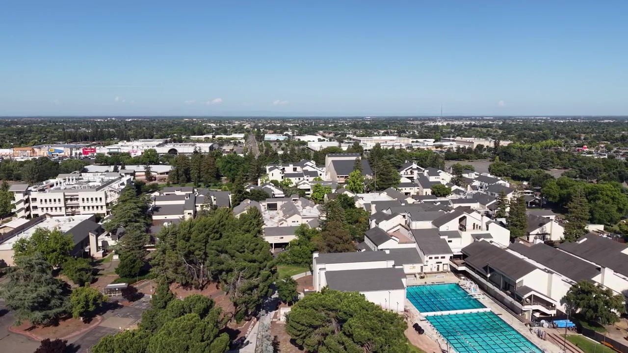 晴れた日のカリフォルニア州ストックトンにて、サンホアキンデルタカレッジのキャンパス（建物群とプール施設を含む）上空を飛行する空撮。背景には市街地が広がる。