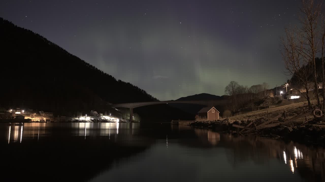 Aurora dances over fjord at night with reflections, bridge view, and glowing houses near Osteroy island