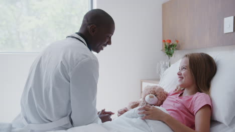 male doctor visiting girl lying in hospital bed hugging teddy bear