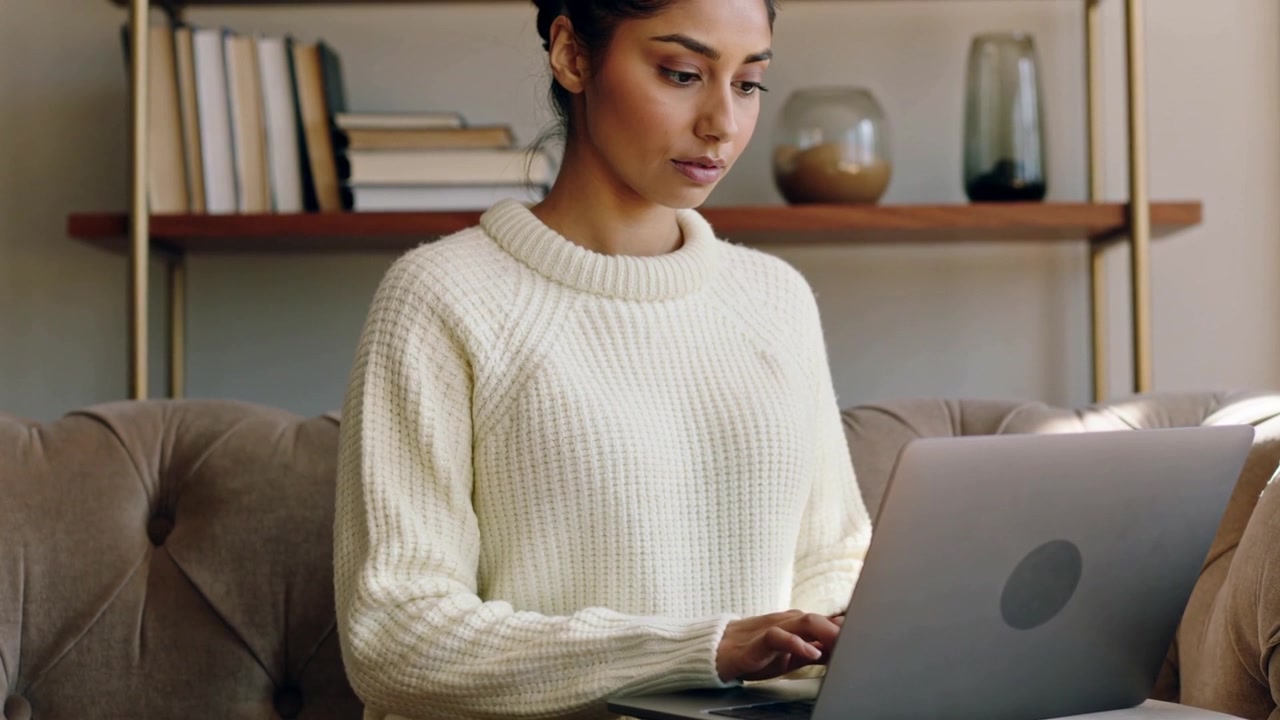 Typing woman wearing cream sweater focusing on laptop on tufted sofa, reading screen