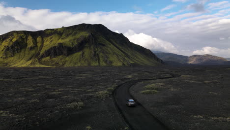aerial push in shot following a car on a black volcanic road with a mountain in the background, on a sunny day with some clouds