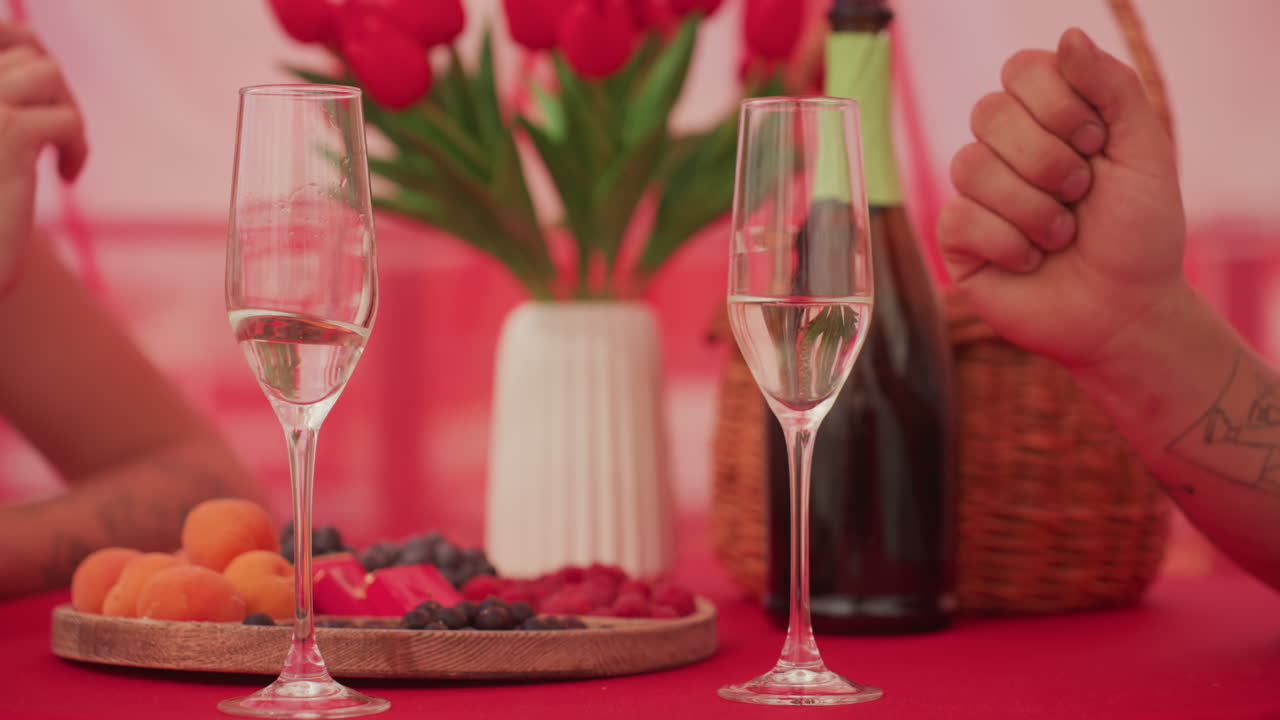 Brindisi intimo durante un picnic con champagne e tulipani. Primo piano delle mani di una coppia che fa tintinnare i flûte sopra una tovaglia rossa, un cesto di vimini, un vaso di tulipani, un piatto di frutti di bosco e albicocche. Una luce calda e soffusa crea
