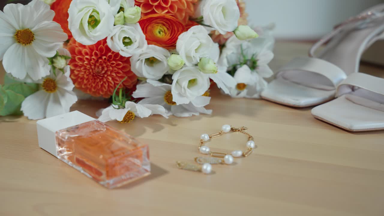 A close-up shot of elegant wedding accessories, featuring white sandals, pearl bracelet, perfume, and vibrant flower bouquet on a wooden floor. Captures timeless bridal elegance