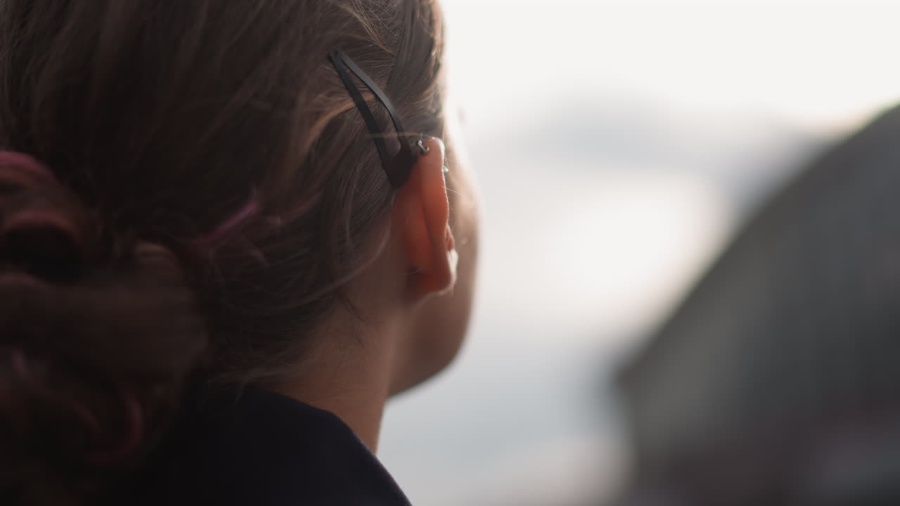 Caucasian woman profile in rainy city, gazing toward blurred skyline, raindrops clinging to braid and ear, soft overcast light, puddle reflections on wet pavement, contemplative slow pan, moody