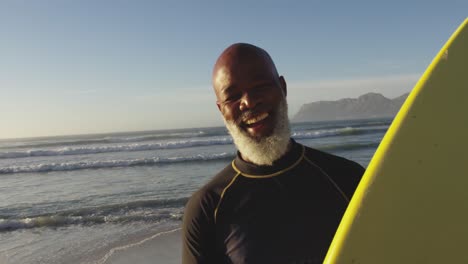 Sonriente-Hombre-Afroamericano-Senior-Caminando-Con-Una-Tabla-De-Surf-En-La-Playa