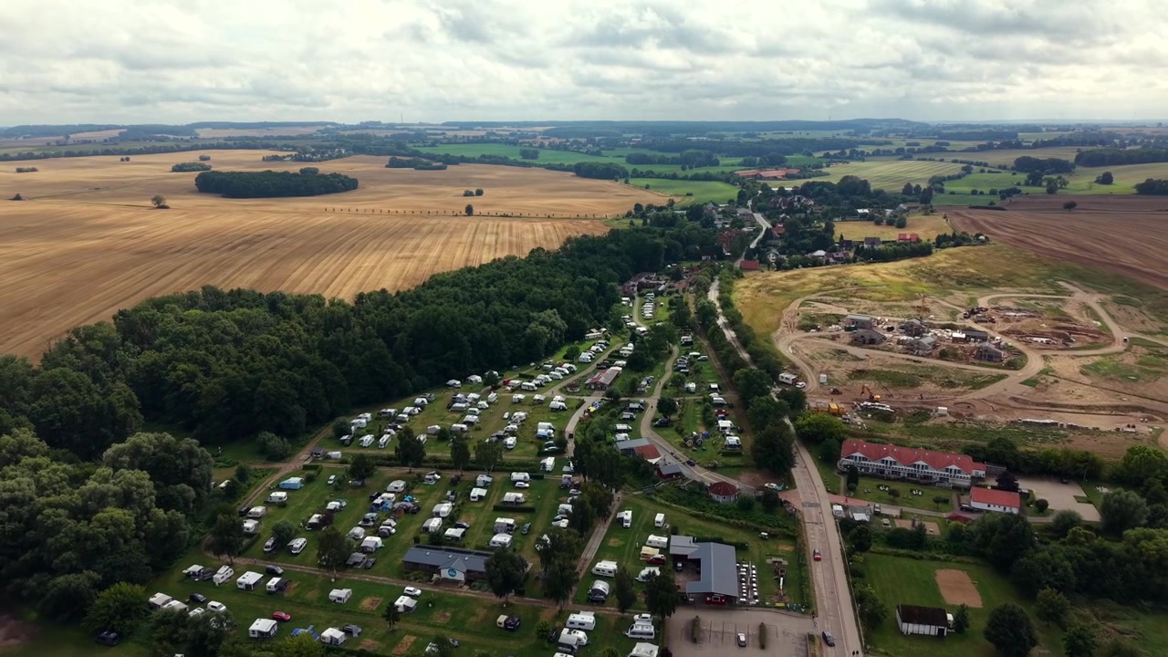 File di roulotte bianche riempiono un campeggio boscoso accanto a campi di grano dorato, un cantiere edile con macchinari pesanti costeggia la tranquilla strada del villaggio vicino al Mar Baltico, Germania. Ripresa aerea con drone in arretramento