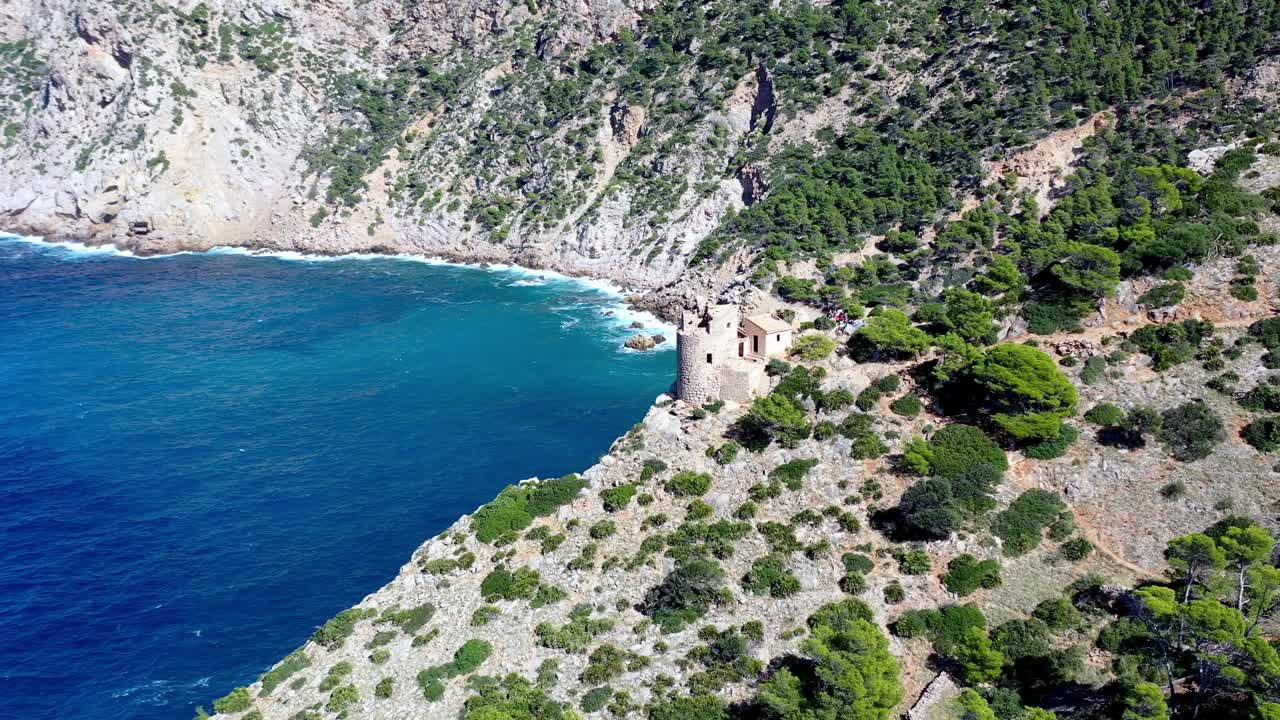 Tower of Basset Cove in Mallorca Spain with tourists exploring and admiring blue waters below, Aerial dolly out reveal shot