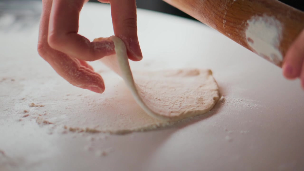 Mani stendono l'impasto con un mattarello di legno su una superficie infarinata. La farina spolvera il piano di lavoro mentre l'impasto si appiattisce. La persona si concentra attentamente, mostrando calma concentrazione. Ideale per ricette, tutorial di cucina.