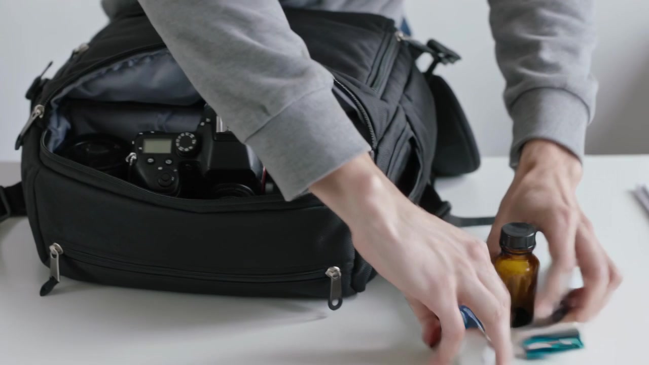 Man in grey sweatshirt placing backpack packing DSLR at white table tucking toiletries for shoot