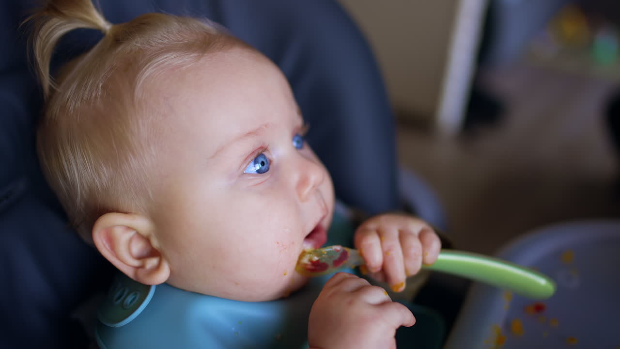 Face of a blond blue-eyed infant with a bib. Little kid is chewing a spoon during meals. Close up.