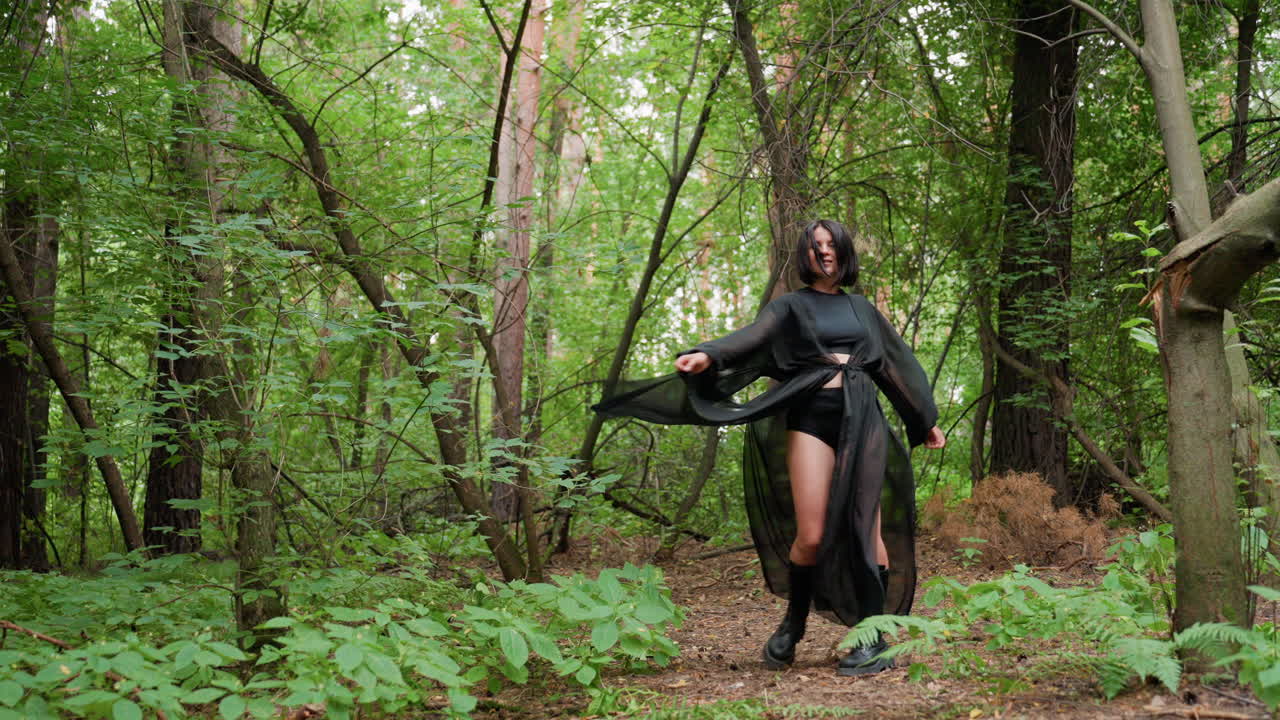 Young girl wearing black sheer outfit and boots turning around gracefully in lush green forest, movement of flowing fabric and soft daylight creating dreamy cinematic atmosphere filled with mystery